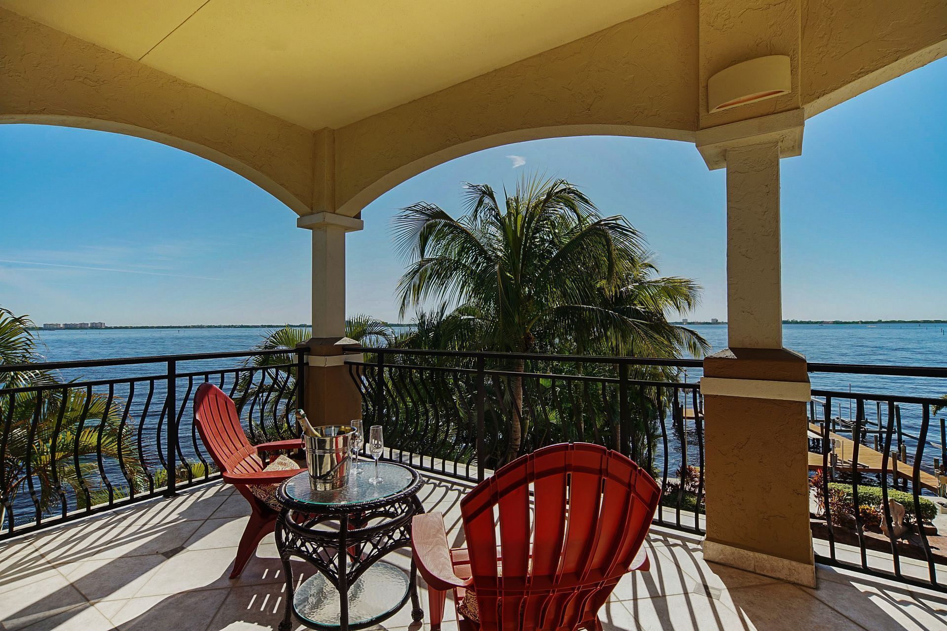 A covered outdoor seating area with a table and chairs, overlooking a body of water and palm trees.