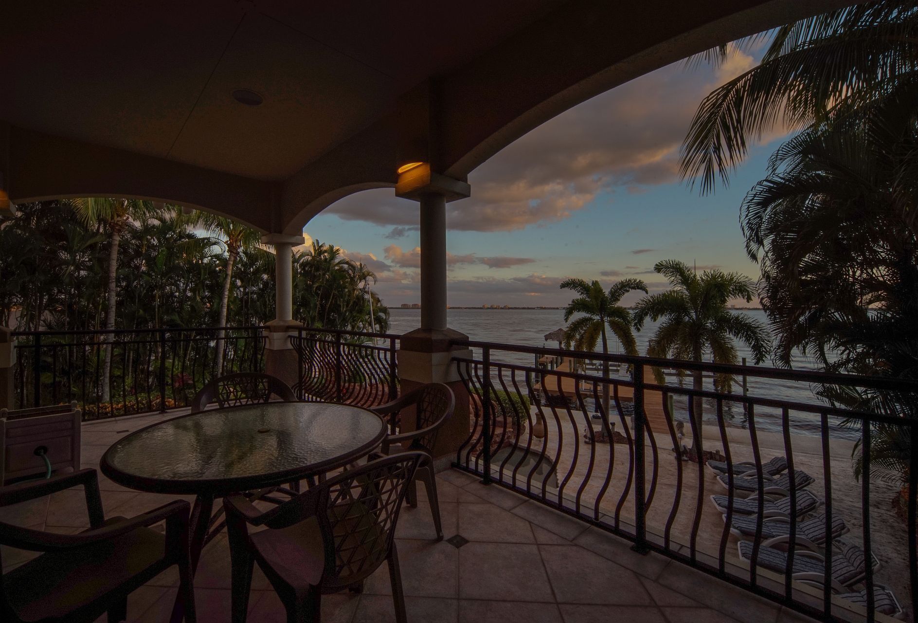 A balcony with a glass table and chairs, overlooking a beach with palm trees and a sunset view.