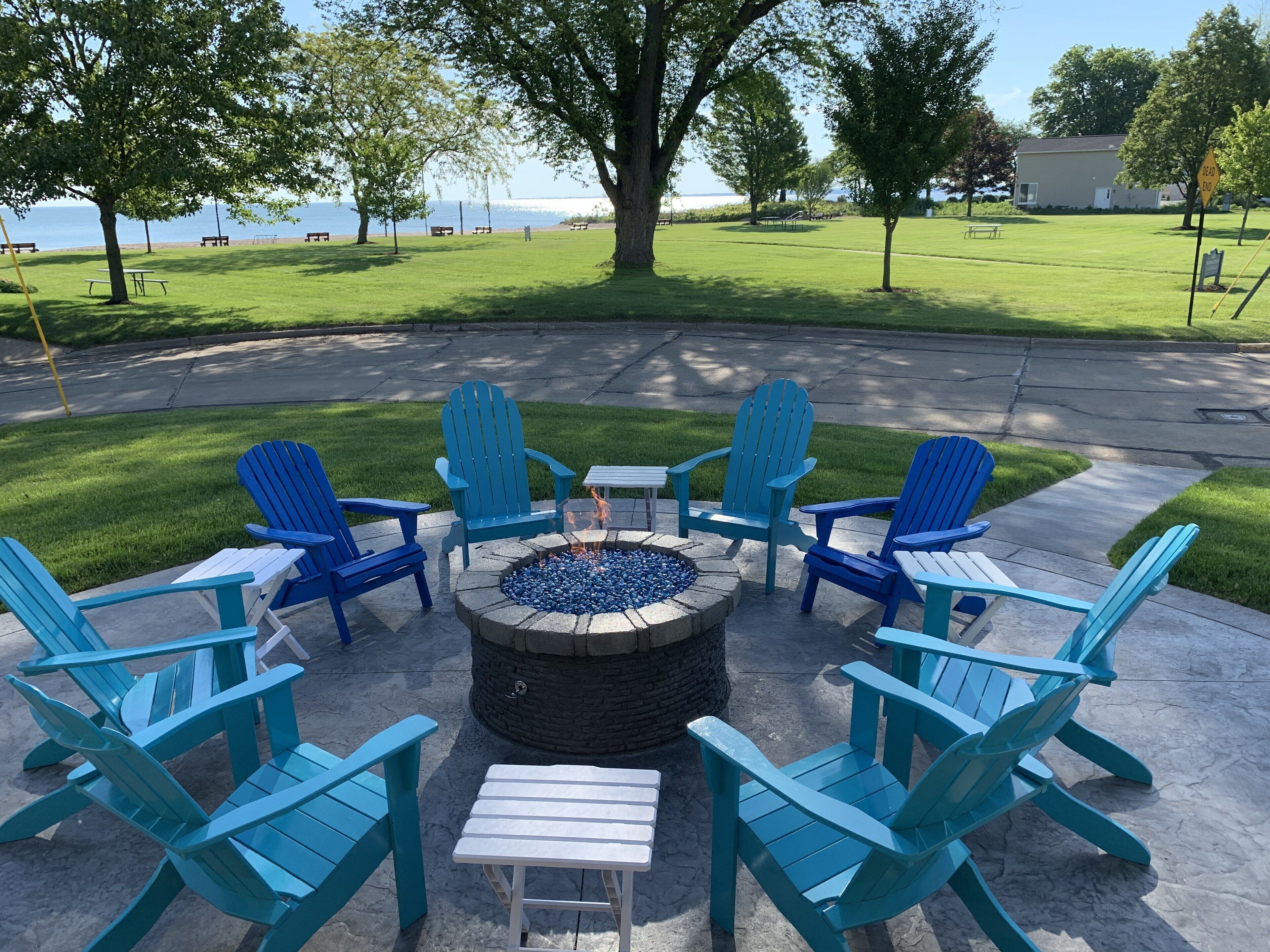 Un fuoco circonda poltrone Adirondack blu e tavoli bianchi su un patio con vista su un'area erbosa e uno specchio d'acqua.