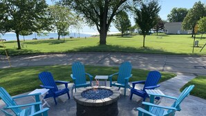 A fire pit surrounded by blue Adirondack chairs and white tables on a patio with a view of a grassy area and a body of water. - OLD HOMESTEAD #1- STUNNING LAKE VIEWS! STEPS FROM LAKE ERIE! (Huron)