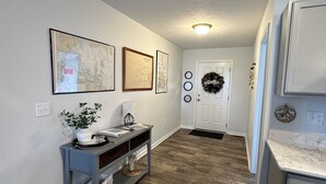 A hallway with a console table, framed artwork, a door with a wreath, and a kitchen area with cabinets. - OLD HOMESTEAD #1- STUNNING LAKE VIEWS! STEPS FROM LAKE ERIE! (Huron)