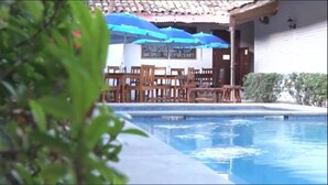 Zona de piscina con sillas y mesas de madera, sombrillas azules y un letrero que indica reservas de habitaciones.