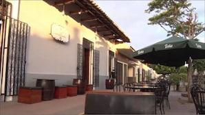 Un área de estar al aire libre con mesas y sillas, un paraguas verde y un edificio con fachada blanca y detalles de metal decorativo.