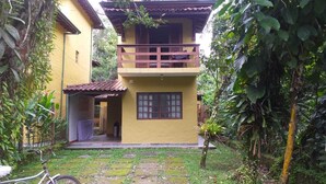 A two-story building with a balcony, surrounded by dense vegetation. - HOUSE IN VILLAGE CAMBURI, R. PRAIA DA MARANDUBA 253, INDIVIDUAL AND ANNUAL LEASES (São Sebastião)