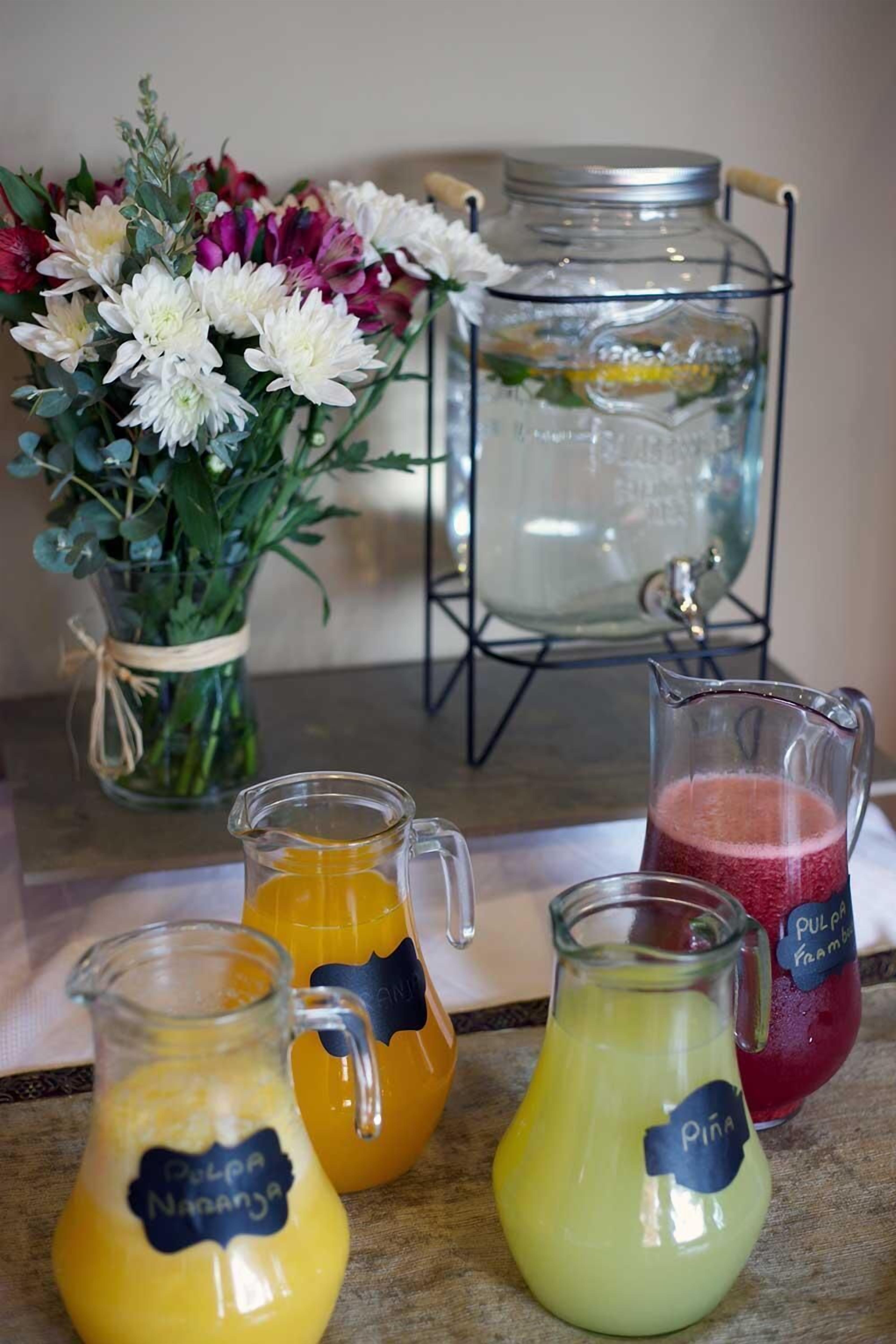 A table with a floral arrangement, a water dispenser, and three labeled pitchers of juice.