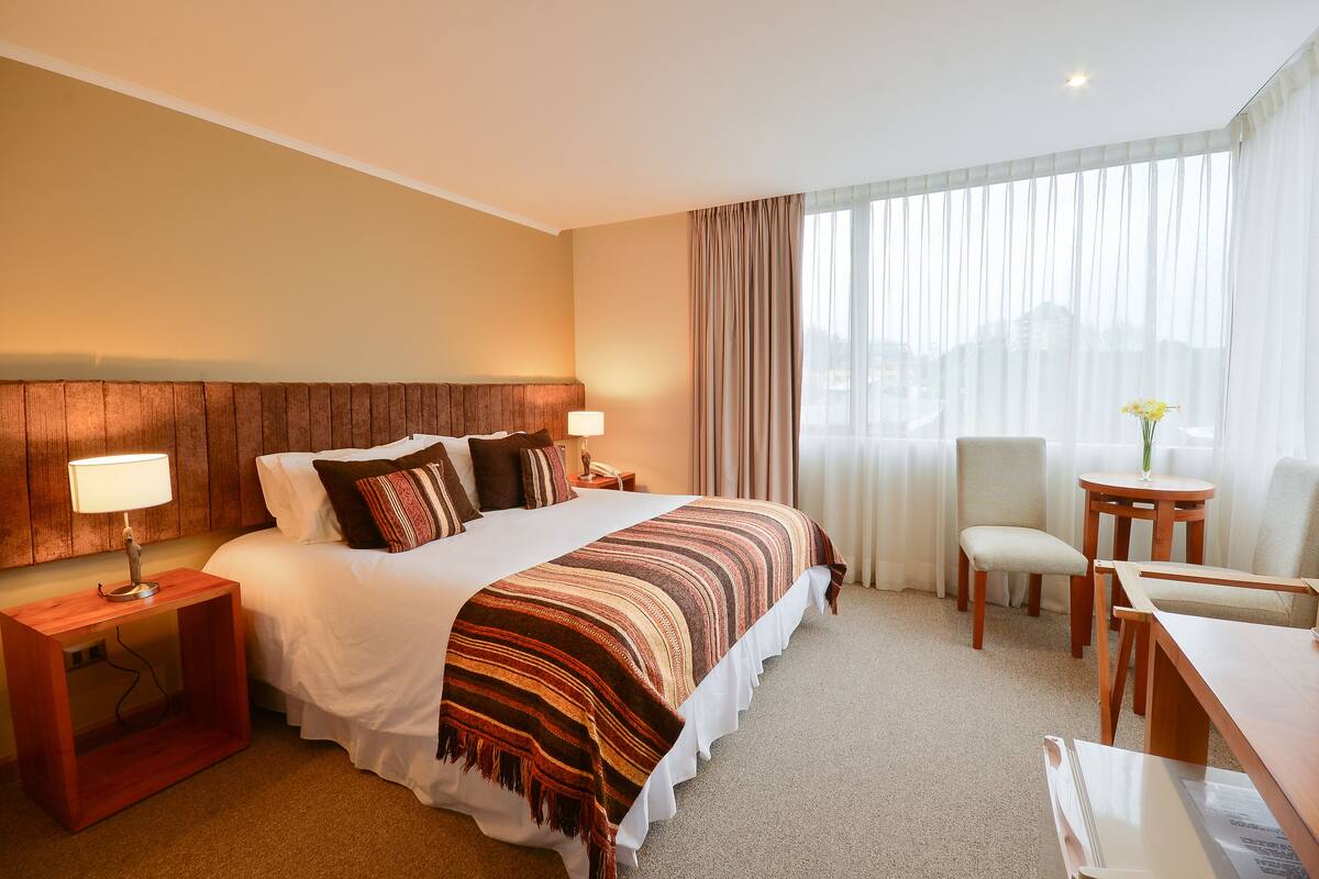 Quarto de hotel com uma cama grande, mesa de cabeceira de madeira, escrivaninha, cadeira e janela com cortinas.