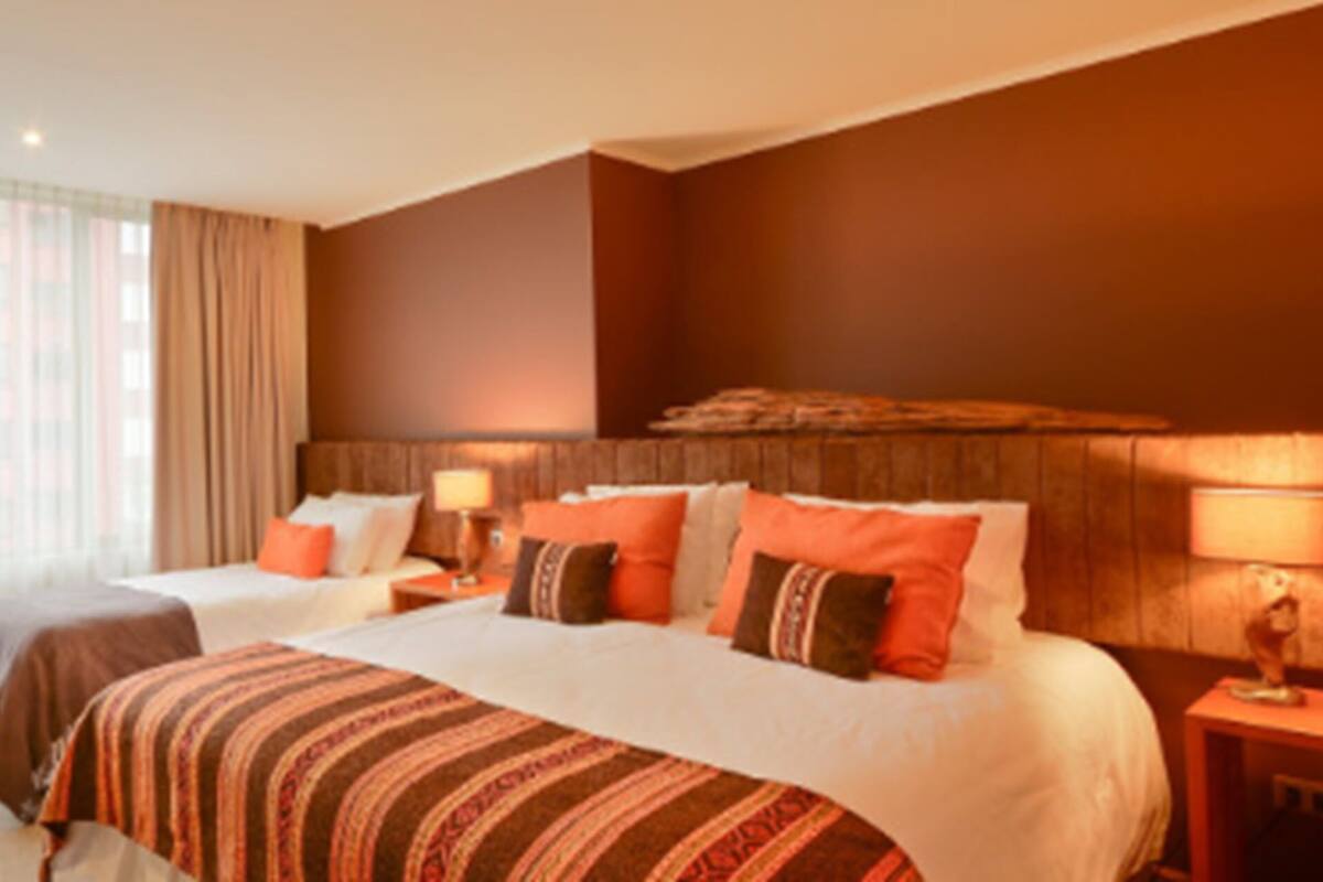 Quarto de hotel com duas camas, uma escrivaninha, uma cadeira e uma janela grande com cortinas.