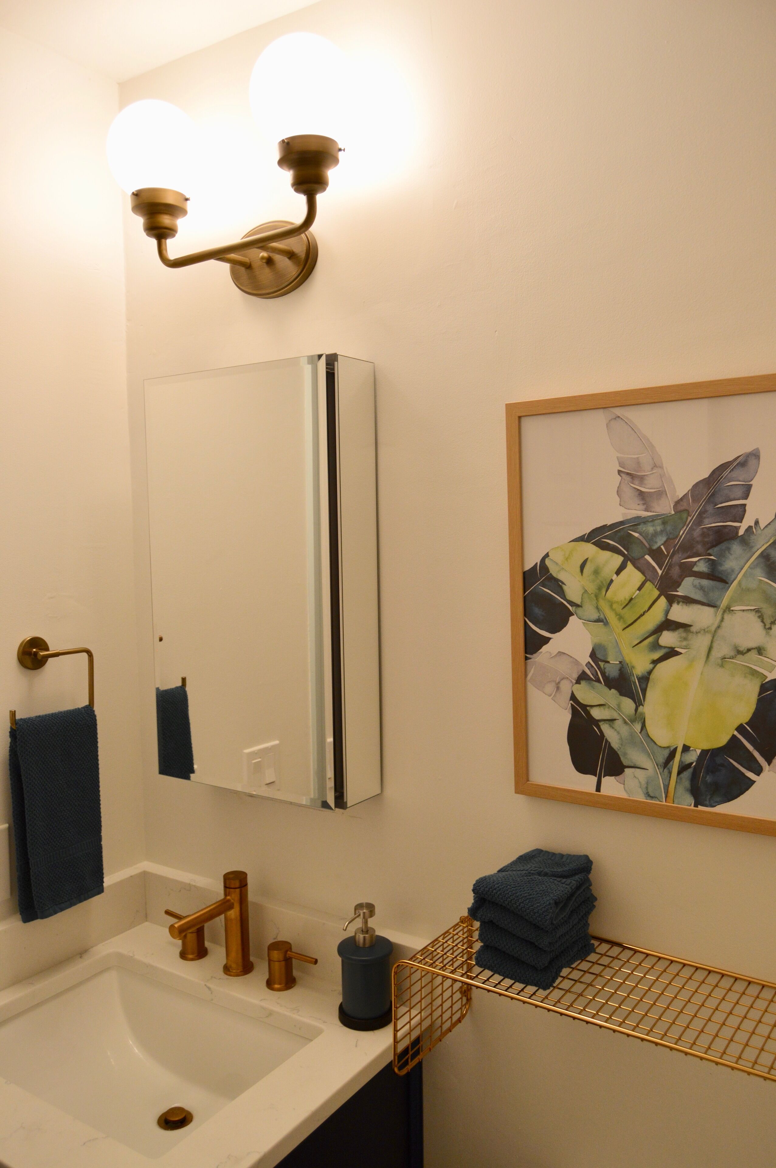 A bathroom with a white sink, gold fixtures, a blue towel, a framed plant, and a mirror.