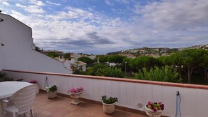 A terrace with a white table and chairs, potted plants, and a view of a cityscape under a partly cloudy sky. - Voramar CostaBravaSi - Pool, BBQ, views, wifi (S'Agaró)