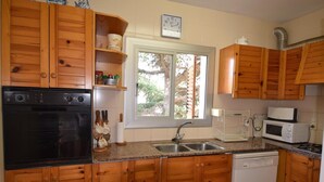 A kitchen with wooden cabinets, a black dishwasher, a white microwave, and a window with a view of trees. - Voramar CostaBravaSi - Pool, BBQ, views, wifi (S'Agaró)
