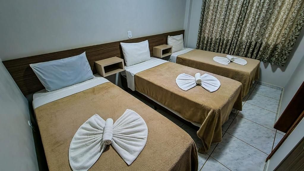 A hotel room with two single beds, each with a brown bedspread and a towel arranged in a bow shape. There are bedside tables with lamps and a curtain with a floral pattern.