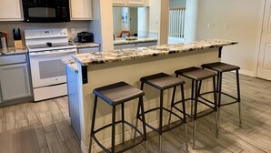 A modern kitchen with white cabinetry, a black microwave, a stove, and a marble countertop. There are three bar stools and a small island with a sink.