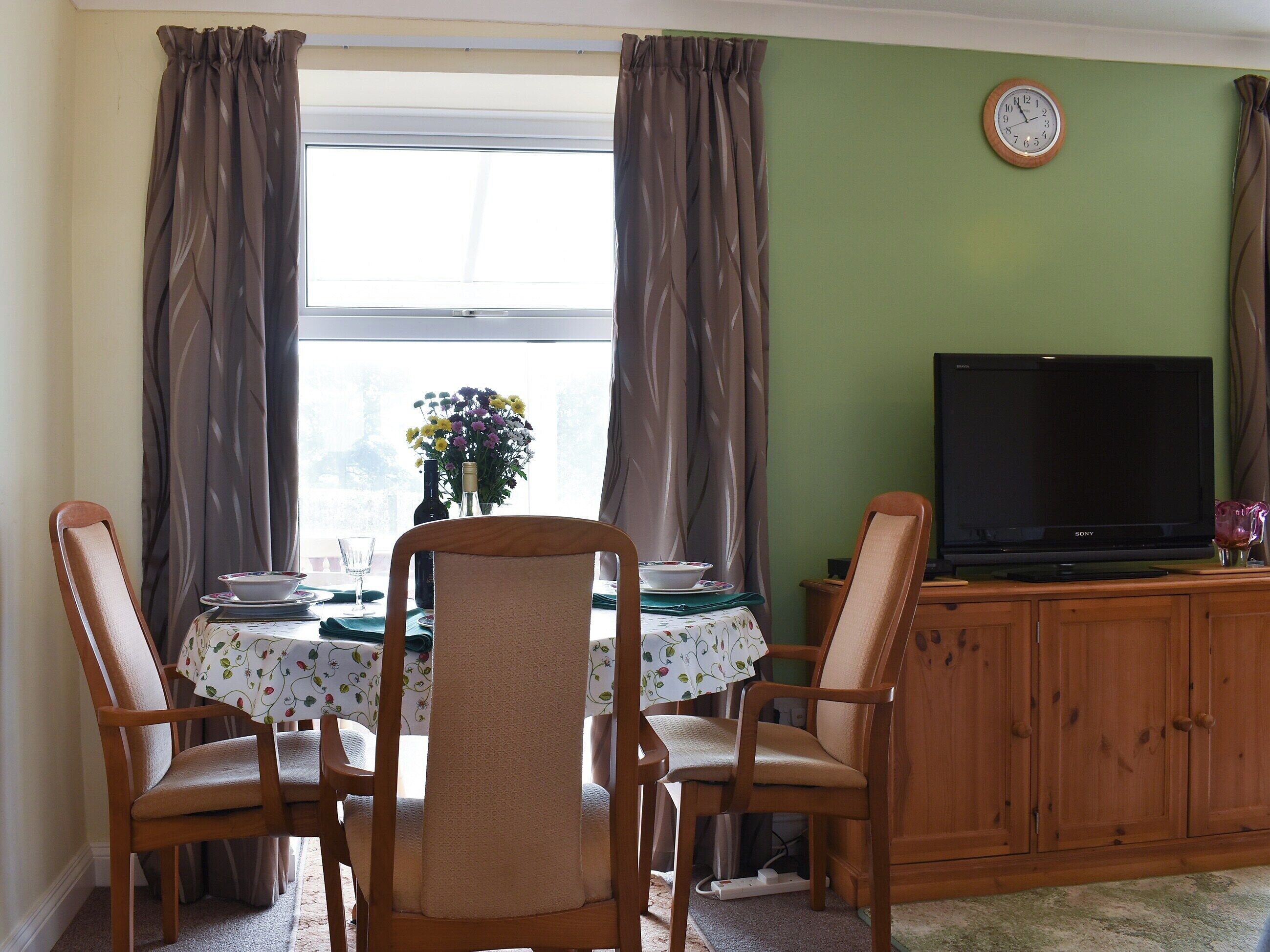 Un comedor con una mesa lista para dos personas, un televisor sobre un mueble de madera y un reloj en la pared.