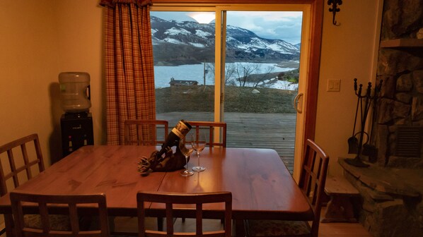 A dining area with a wooden table, four chairs, a bottle of wine, and two wine glasses. A water dispenser is on the table, and a view of snow-capped mountains is visible through the window.