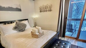 A modern bedroom with a large bed, a bench, a view of trees through a glass door, and a wall-mounted light fixture.
