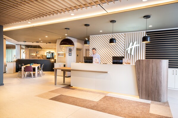 Una recepción moderna con un mostrador de madera, un área de estar con una mesa y sillas, y un muro decorado con un diseño.