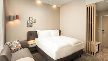 A hotel room with a bed, a nightstand, a lamp, a sofa, and a wall-mounted shelf.