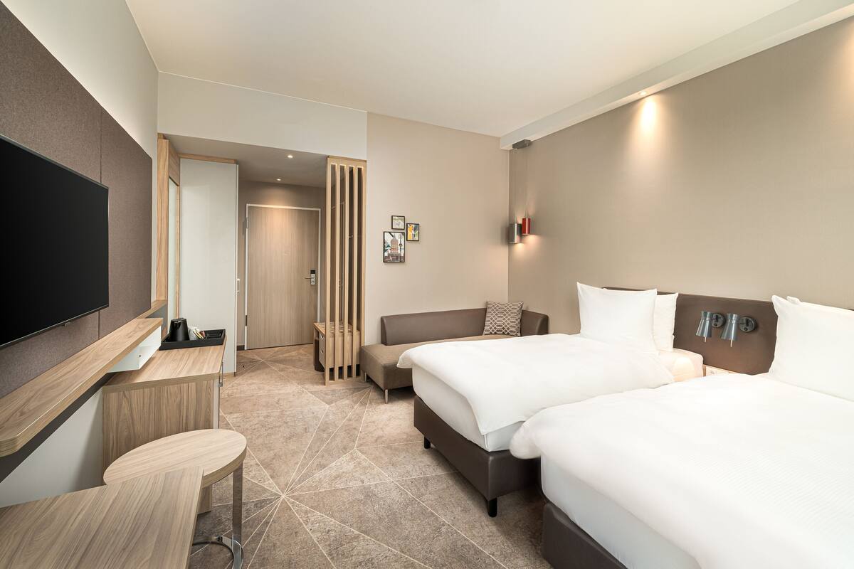 Ett modernt hotellrum med en platt-TV, ett skrivbord, två sängar, en soffa och en väggmonterad lampa.