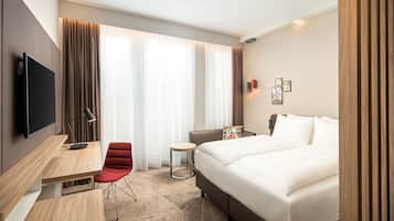 A modern hotel room with a large bed, a desk with a chair, a small round table, and a TV mounted on the wall.