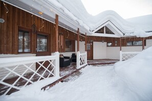 A snow-covered wooden cabin with a porch, a covered outdoor seating area, and a view of the snowy landscape.