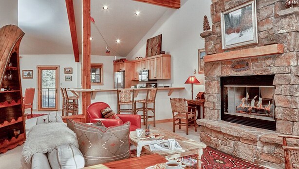 Un salon chaleureux avec une cheminée en pierre, des poutres en bois et un coin repas agrémenté d’une chaise rouge.