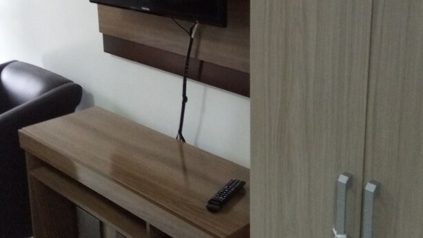 A room with a wooden TV stand, a mounted flat-screen TV, a remote control, and a closed wardrobe. - Studio apartment in a condominium with air conditioning. (Curitiba)
