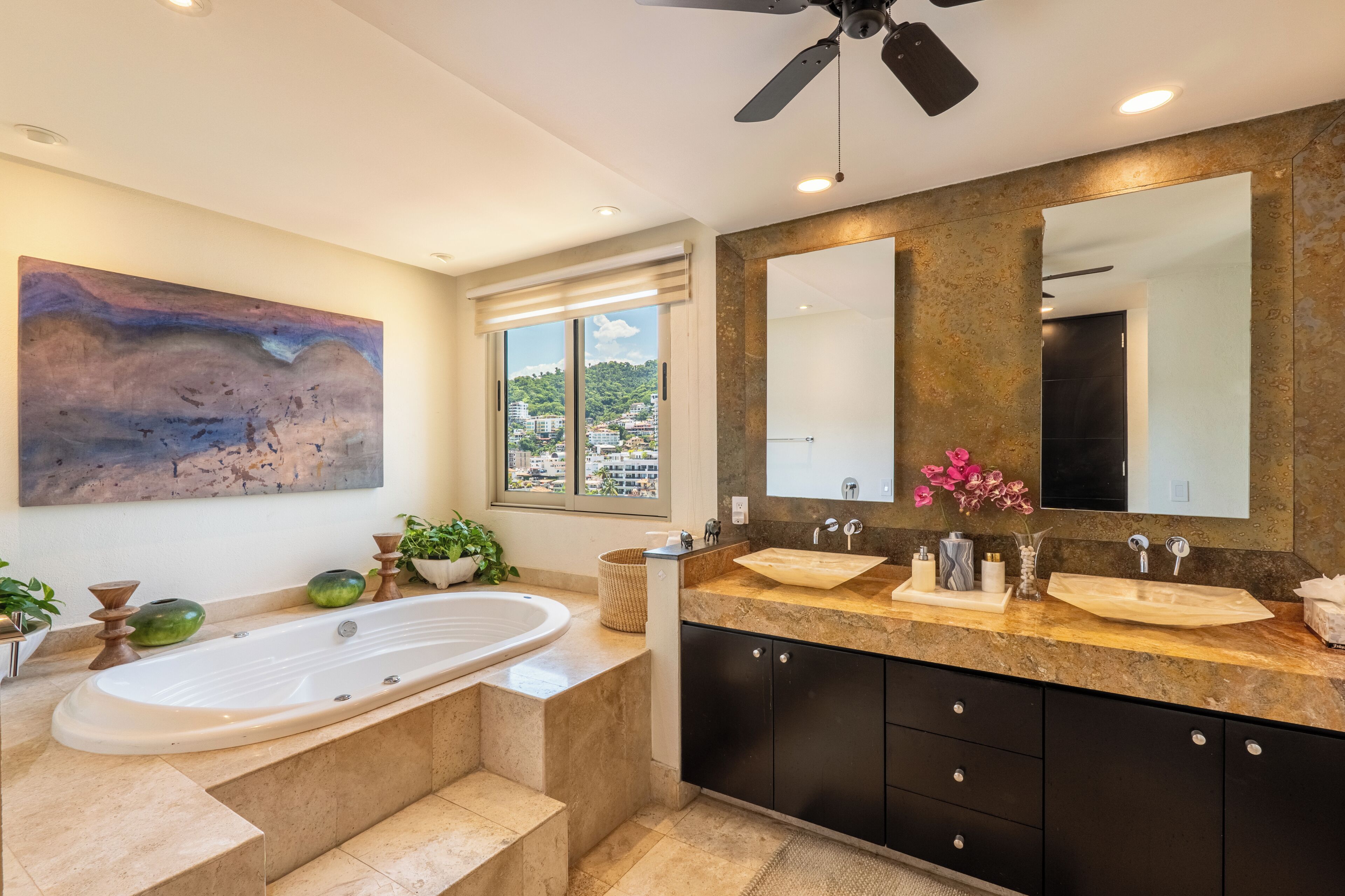 Un bagno moderno con una vasca da bagno ampia, due lavandini, una ventola a soffitto e una grande finestra con vista sulla città.