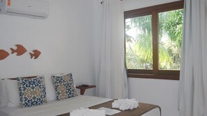 A hotel room with a bed, patterned pillows, a wooden headboard, a window with curtains, and an air conditioning unit. - Bahia Bonita Flat (Itacaré)