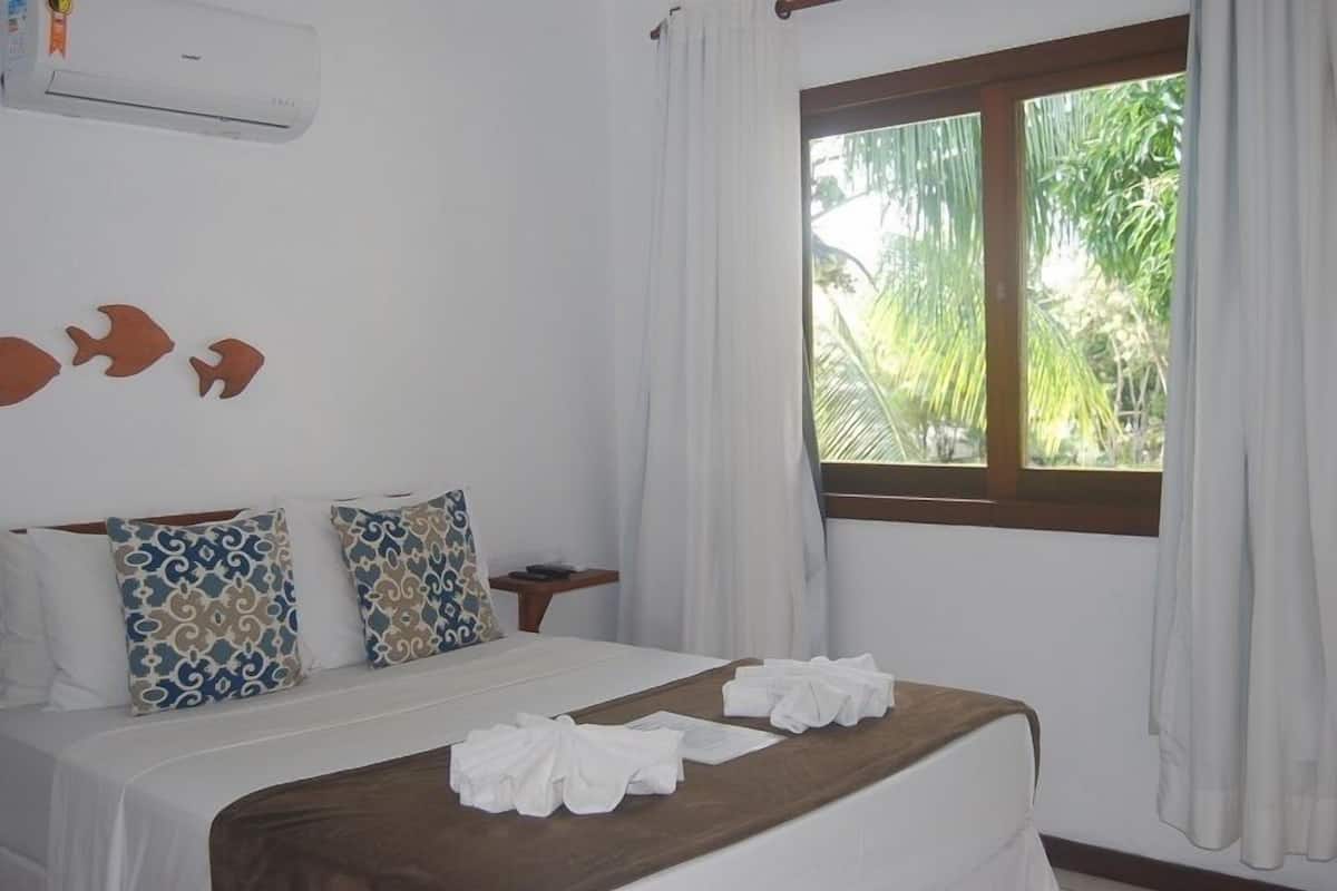 Une chambre d’hôtel avec un lit, des coussins à motifs, une tête de lit en bois, une fenêtre avec des rideaux et un appareil de climatisation.