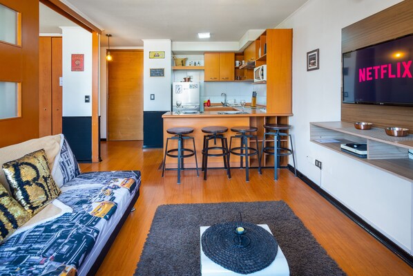A compact living space with a kitchenette, a sofa with patterned cushions, a coffee table, and a TV mounted on the wall. - Perfect Apartmen For Lover Of Travels. Best Located, Full Equiped, Include A/C! (Santiago)
