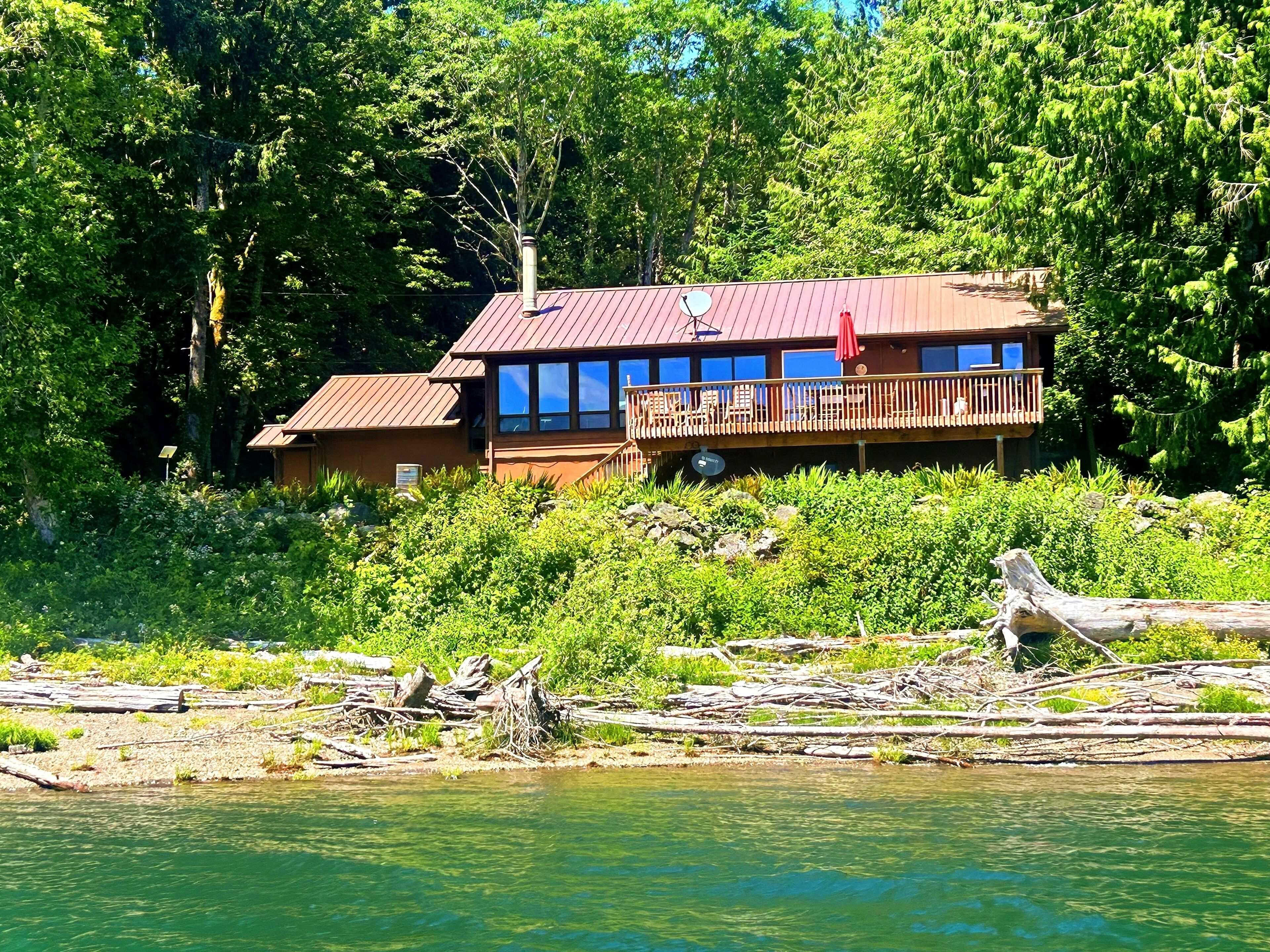 A cabin with a deck, surrounded by dense forest and a body of water.