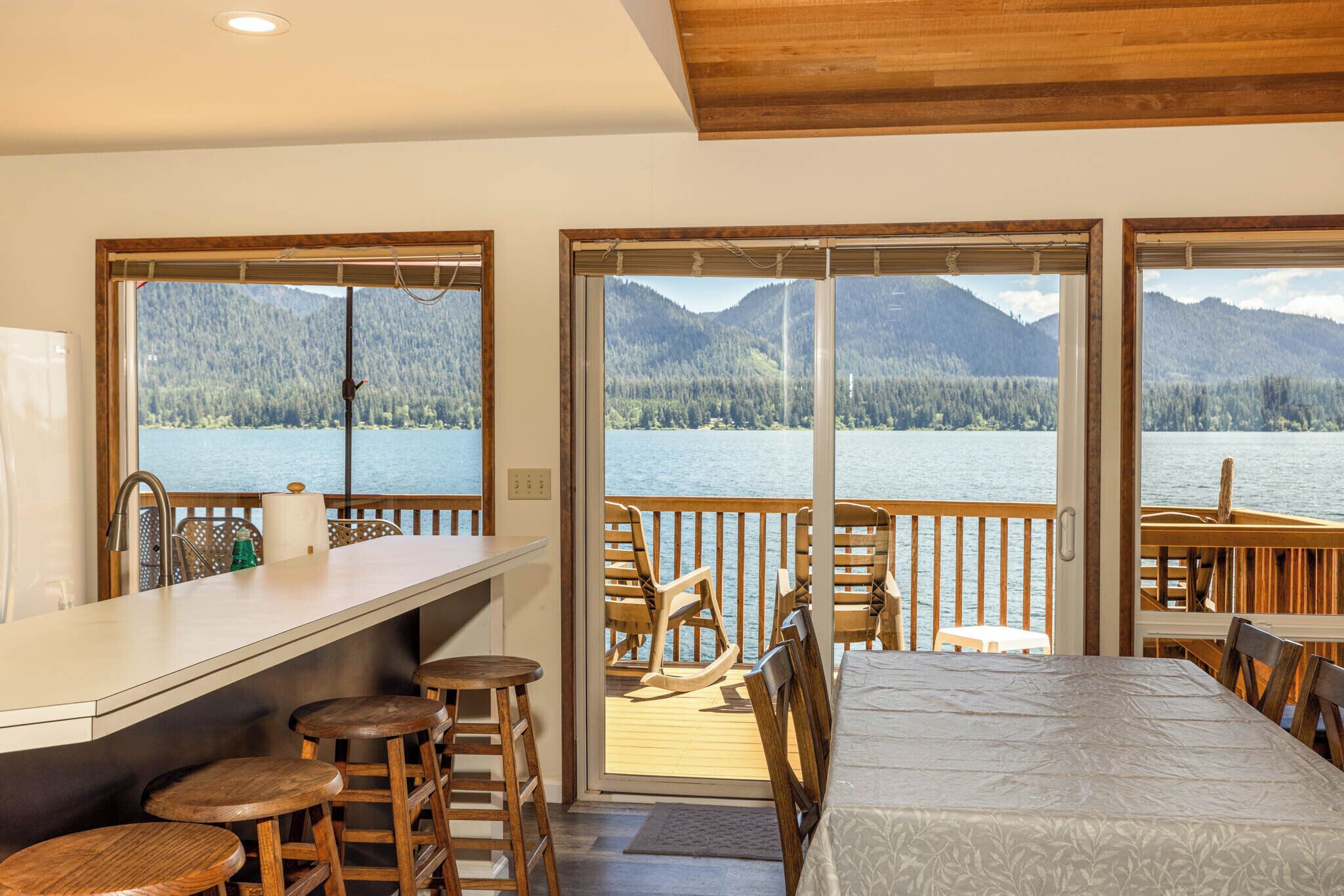 A room with a kitchen counter, bar stools, a dining table, and a view of a lake and mountains through large windows.