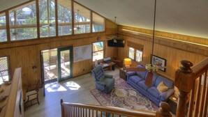 A spacious living room with a high ceiling, wooden walls, and large windows. There is a staircase leading to an upper level, a blue sofa, a green armchair, and a wooden coffee table. - Sound View, Pet Friendly Home in Harkers Island, NC (Core Sound Lodge) (Harkers Island)