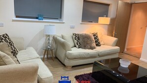 A modern living room with a sofa set, a glass coffee table, and a lamp. There are two windows with blinds, a door, and a logo for 'Beds Away' and 'Airbnb'.