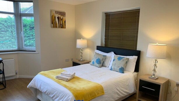 A neatly made bed with a yellow blanket, two bedside lamps, and a window with blinds.