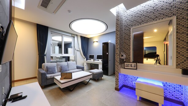 A modern living room with a sofa, coffee table, and a TV. There is a desk with a computer and a mirror. The room has a tiled backsplash and a ceiling light. - Urban Stay (Gwangju)