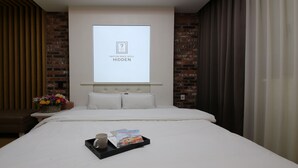 A hotel room with a bed, a projector screen displaying the hotel's name, a bedside table with a vase of flowers, and a tray with a mug and a brochure. - Urban Stay (Gwangju)