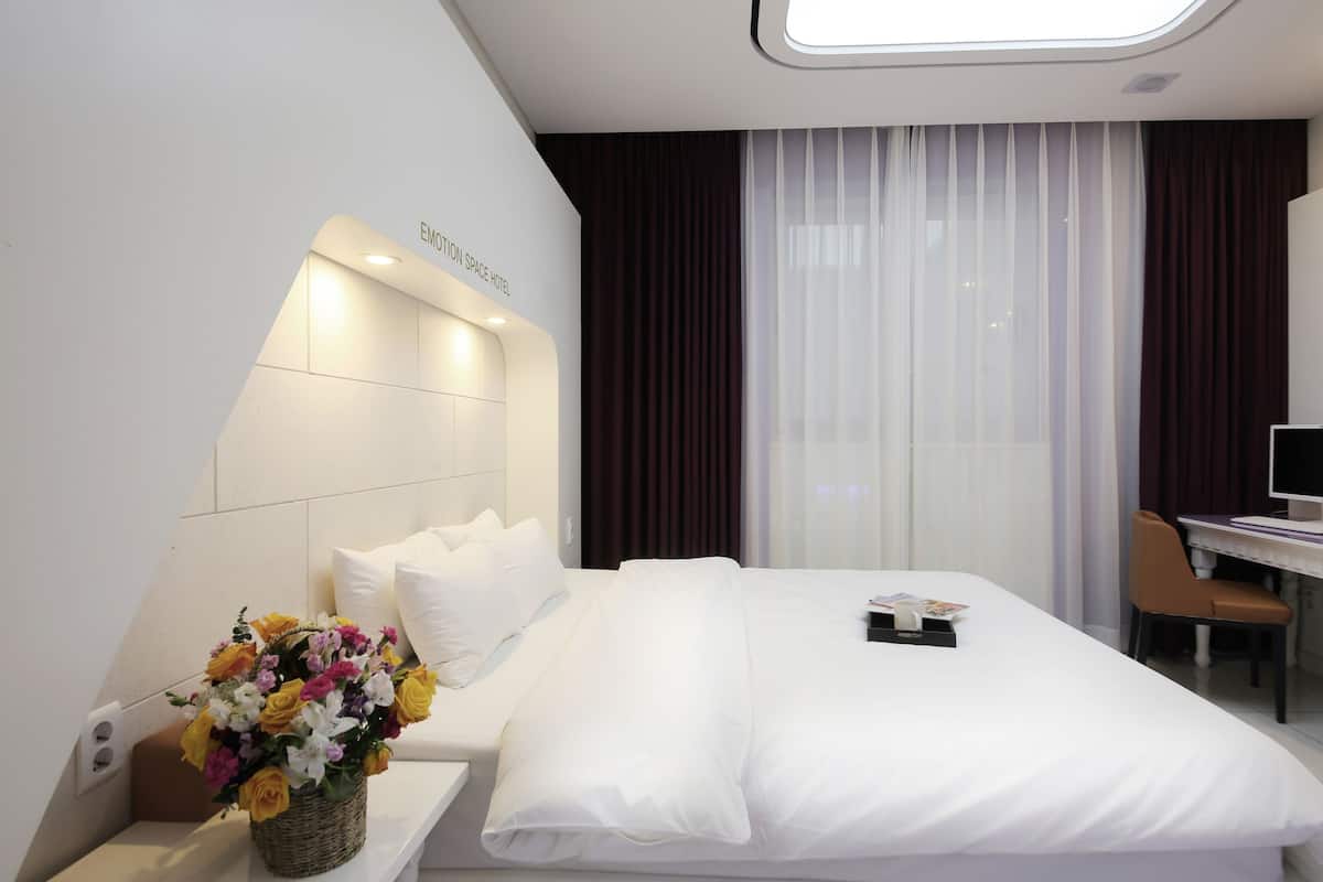 Habitación de hotel con una cama bien hecha, un escritorio con silla, un ventanal con cortinas y un televisor montado en la pared.