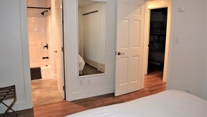 A bedroom with a large bed, a wooden floor, a white door, and a bathroom visible through an open door. - Super Comfy 3 Bed Home, Lovely Patio and Yard with Upscale Seating and Beautiful (Seaside)