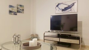 Ein modernes Wohnzimmer mit einem Flachbildfernseher auf einem Standfuß, einem Glastisch mit Kerzen und abstrakter Wandkunst.