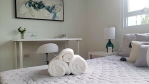 Ein ordentlich bezogenes Bett mit aufgerollten Handtüchern, ein Nachttisch mit Lampe, eine kleine Ablagefläche mit Dekoration und ein Gemälde an der Wand.