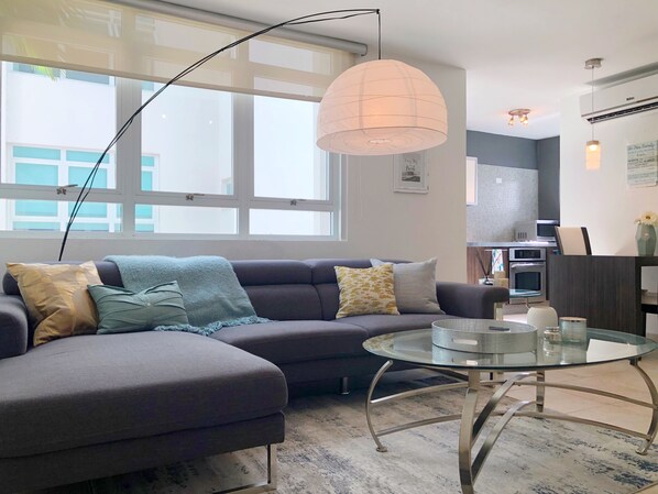 Een moderne woonkamer met een hoekbank, een glazen salontafel en een groot raam dat zorgt voor natuurlijk licht.