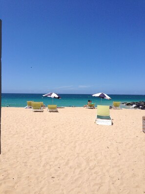 Een strand met meerdere ligbedden en parasols, een heldere blauwe lucht en de oceaan op de achtergrond.