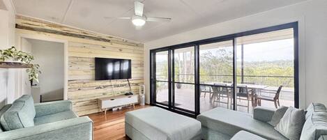 Ampla sala de estar com TV grande, painéis de madeira, ventilador de teto e varanda com área de estar externa.