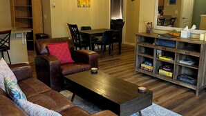Una sala de estar con un sofá de cuero marrón, una mesa de centro de madera, una zona de comedor con mesa y sillas, una estantería con diversos objetos y un espejo grande en la pared.
