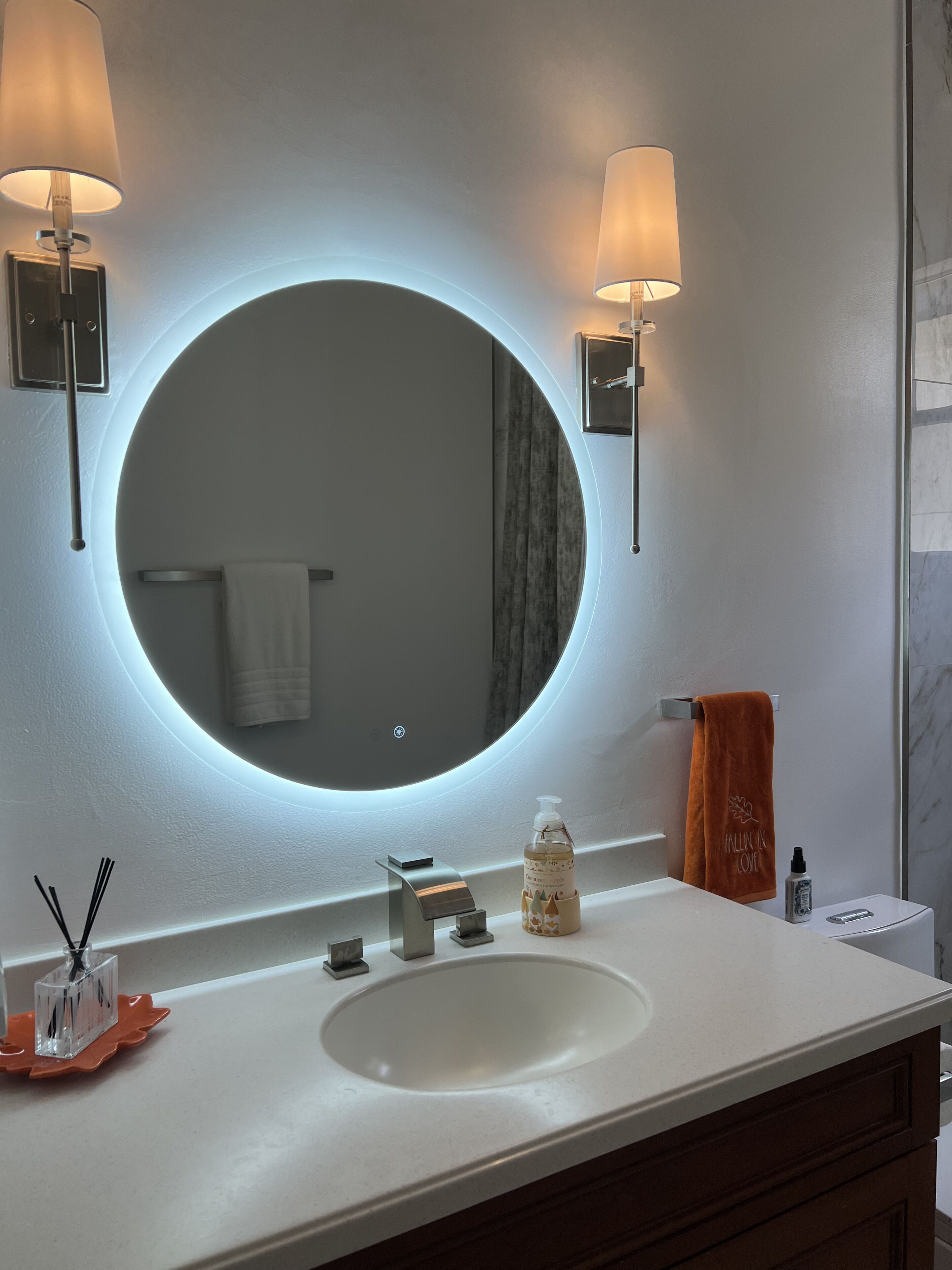 A bathroom with a round mirror, two wall-mounted lights, a white sink, and a towel rack.