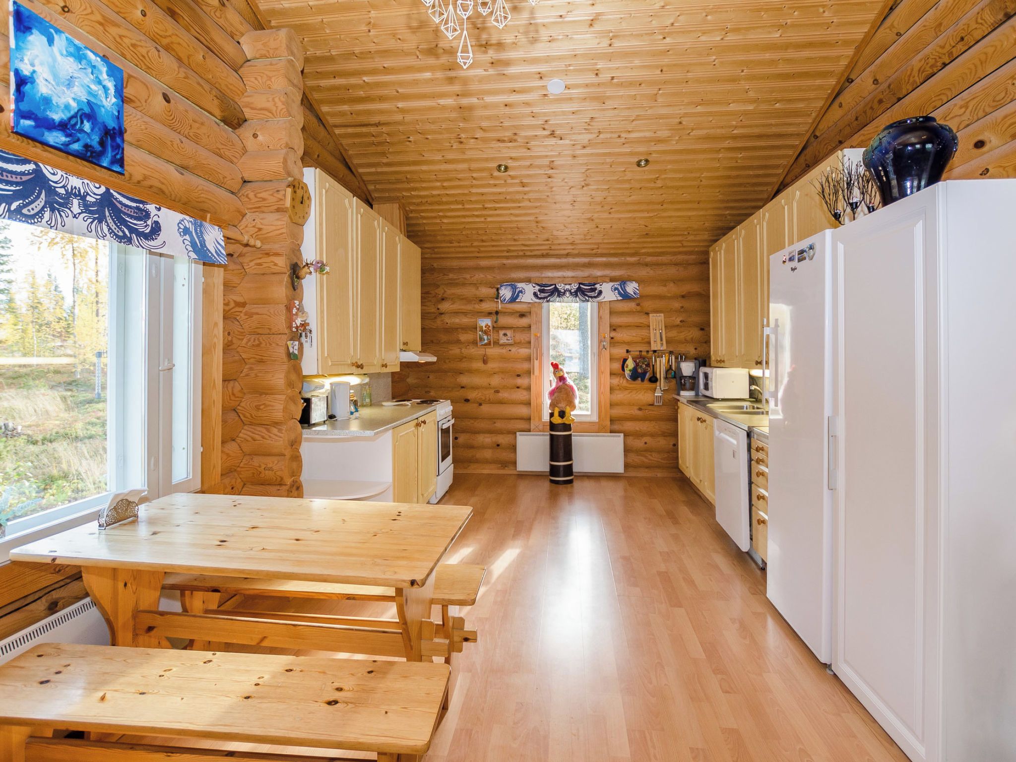 A log cabin interior with a wooden dining table, white cabinets, and a view of the outdoors.
