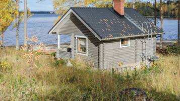 A small cabin with a sloped roof, a chimney, and a window, situated by a body of water with trees in the background.