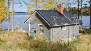 Une petite cabane en bois, dotée d’un toit en pente et d’une cheminée, située au bord d’un lac, avec des arbres en arrière-plan.