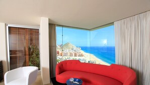 Una moderna camera d'albergo con un divano rosso, una poltrona bianca e un'ampia finestra con vista sulla spiaggia e sullo skyline della città.